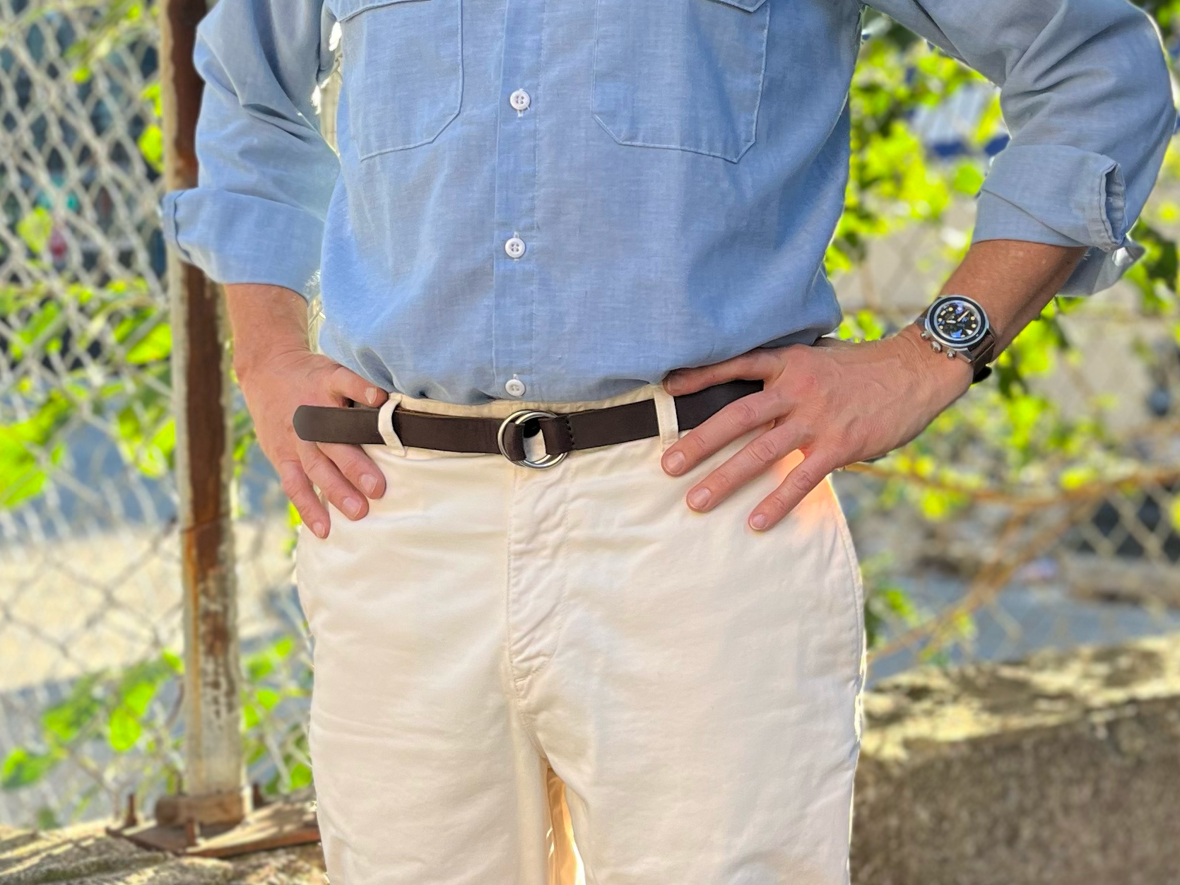 A person stands outdoors with hands on hips, wearing a light blue button-up shirt, cream-colored pants, a brown belt, and a wristwatch. Sunlight and a chain-link fence are visible in the background.