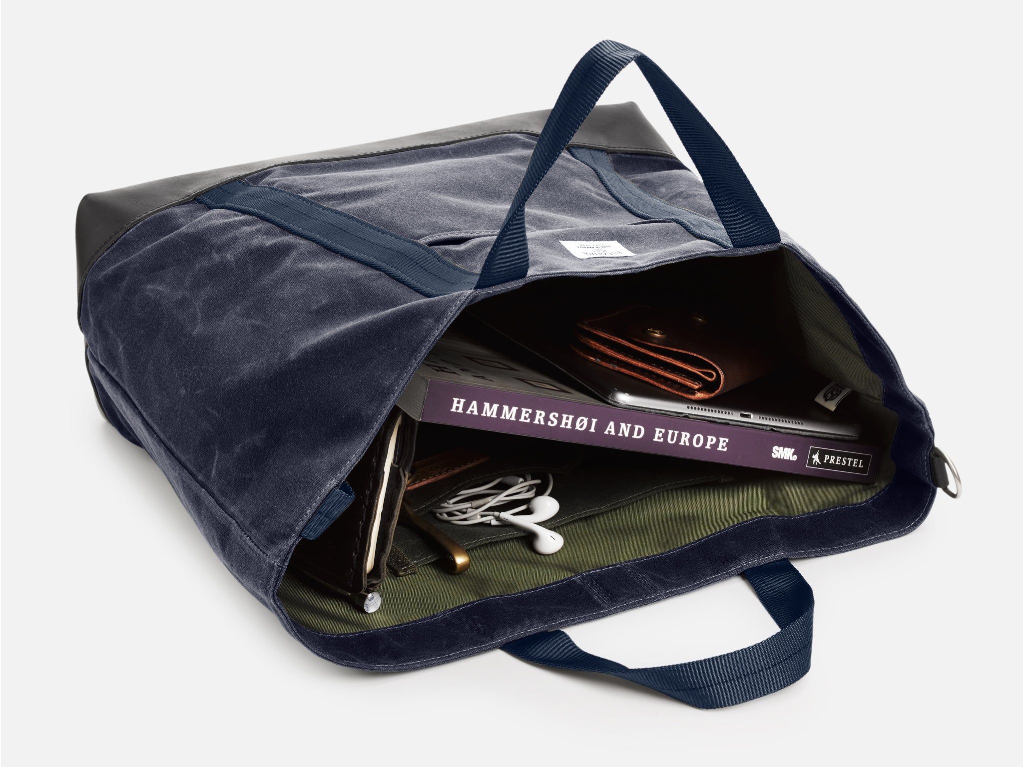 A navy blue tote bag, resting on its back, partially opened, reveals a book titled Hammershoi and Europe, a brown wallet, a tablet, and white earphones neatly inside.