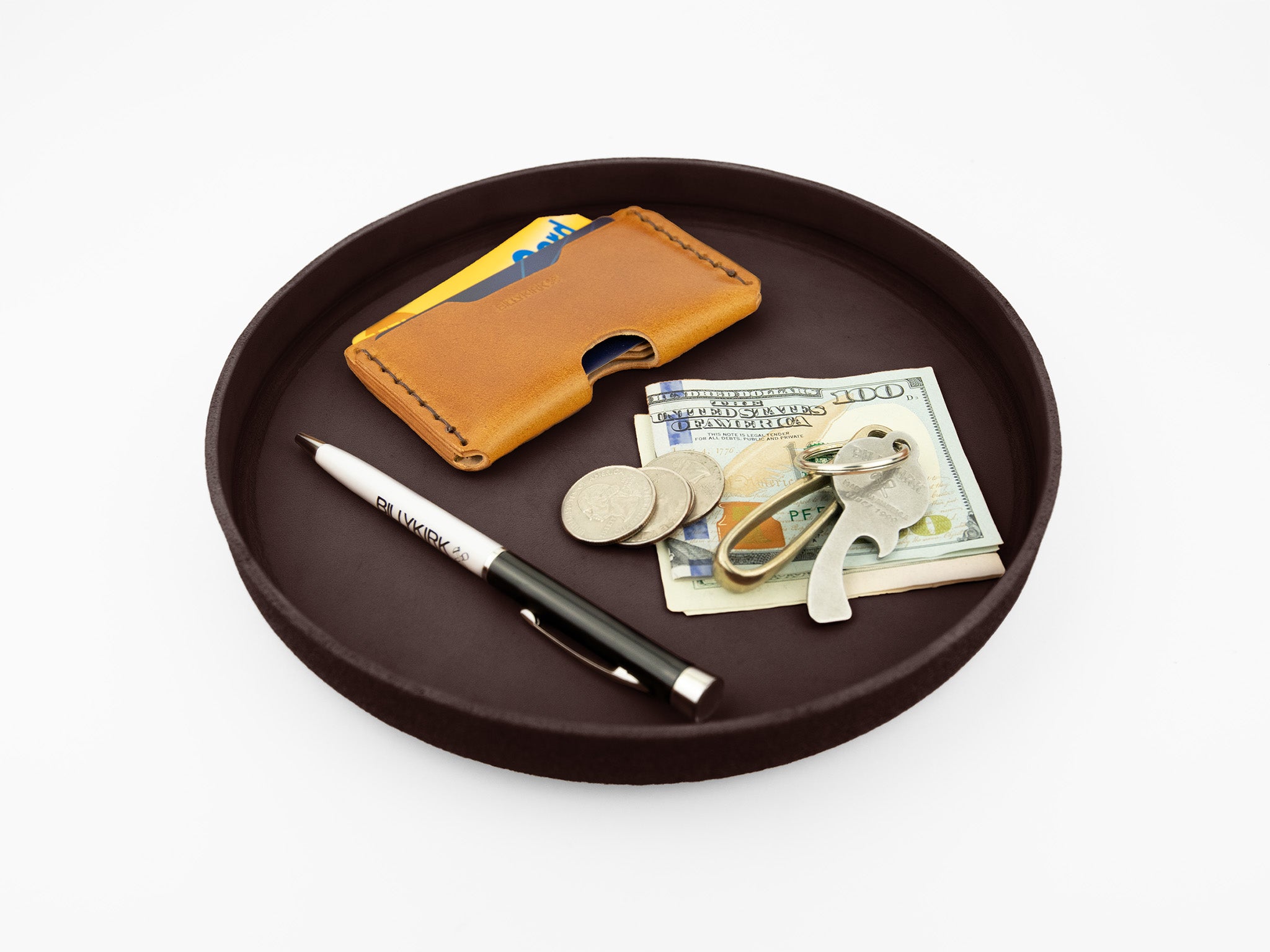 A round black tray holds a tan leather card holder with cards, a folded hundred-dollar bill with coins and keys on top, and a black pen.