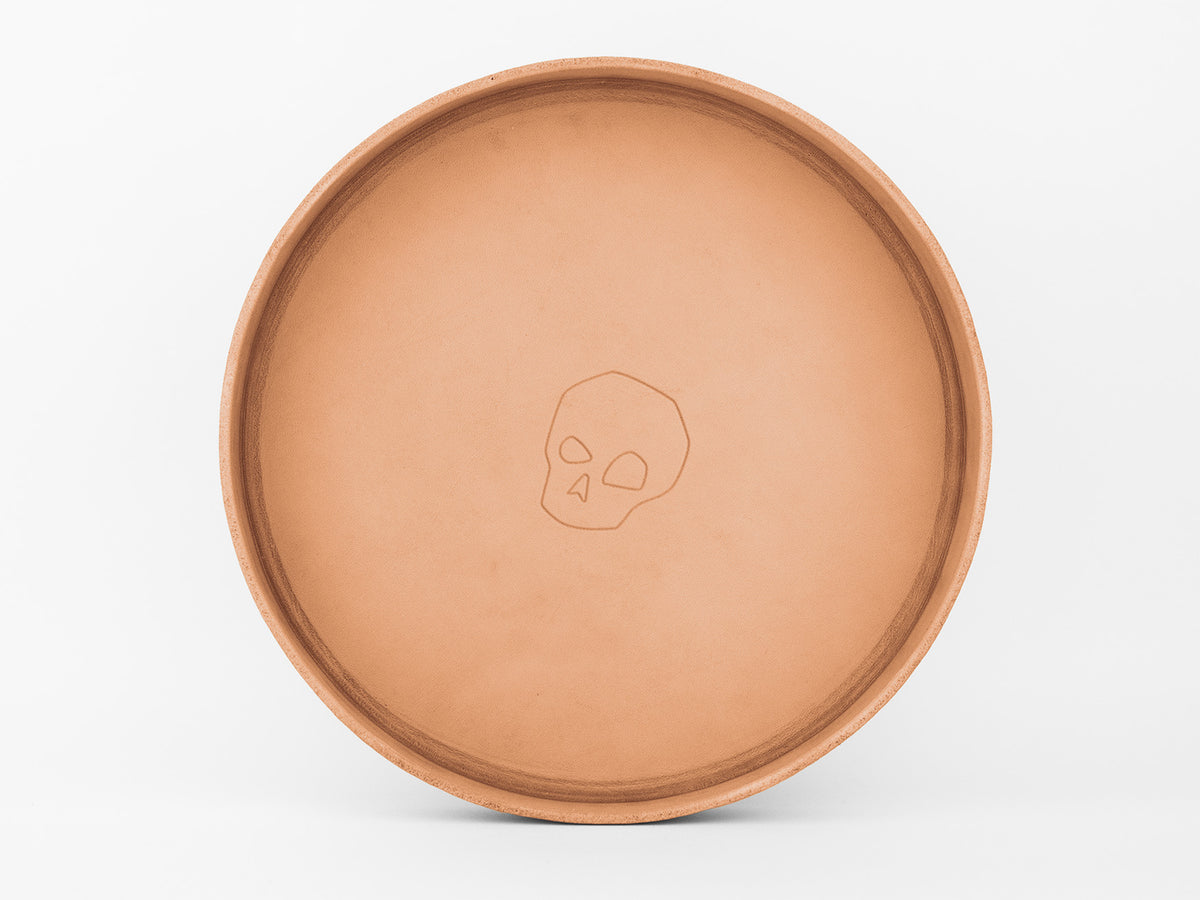 A round, light brown leather tray with a simple, embossed skull design in the center, placed against a white background.