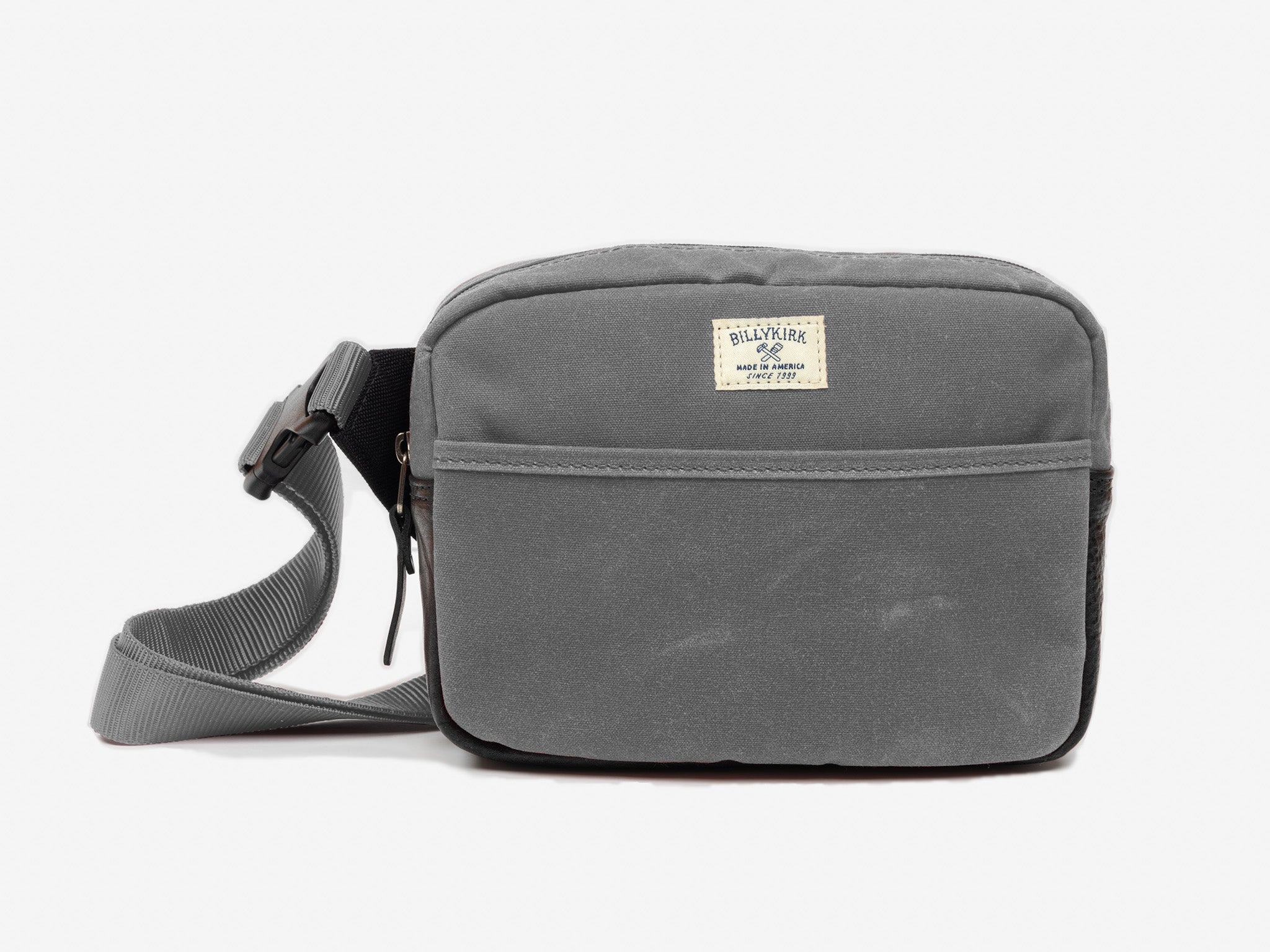 A small ash waxed canvas crossbody bag with a front pocket, adjustable strap, and a cream colored rectangular Billykirk brand patch stitched on the front.