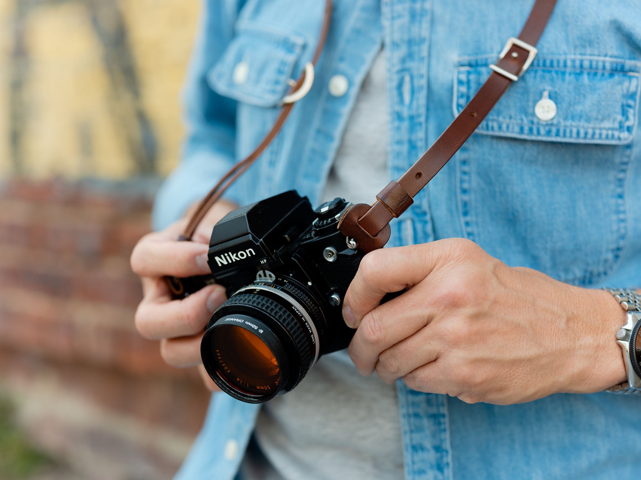 A person wearing a denim shirt holds a Nikon film camera with both hands, the camera hanging from a brown leather strap around their neck.