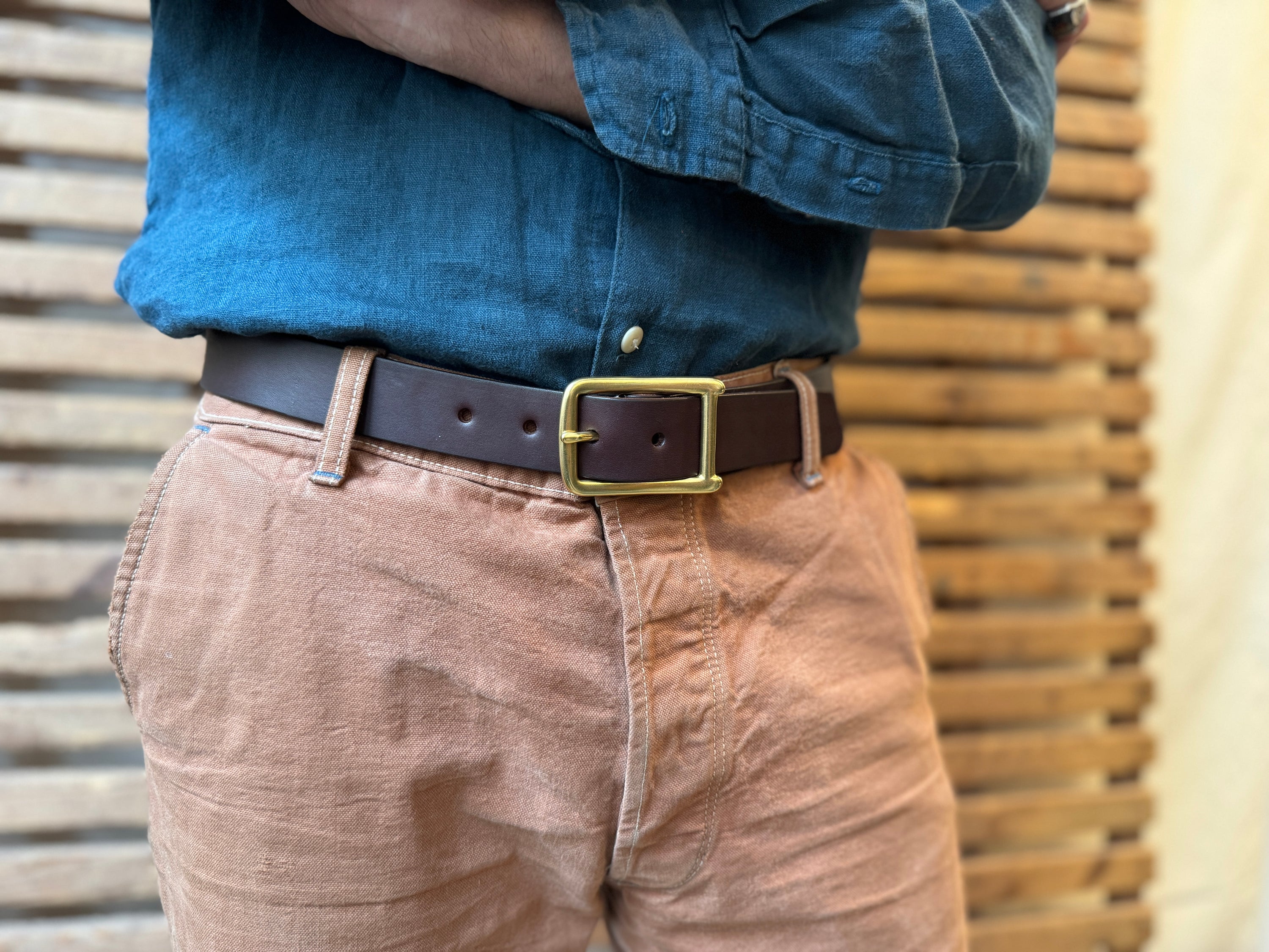 A person wearing a blue denim shirt tucked into light brown pants with a dark brown leather belt and brass buckle, standing in front of a wooden slat background.