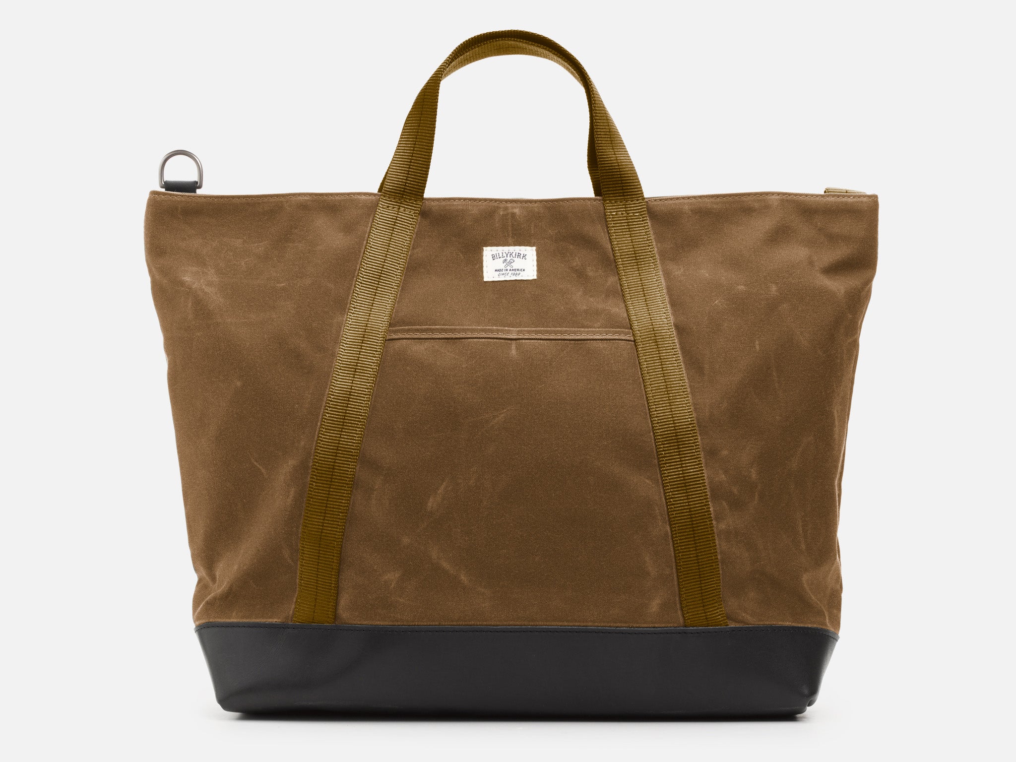A front view of a brush brown waxed canvas tote bag with 1" nylon brush brown handles and shoulder strap, a small off-white label on the front, and a black leather bottom, set against a plain white background.