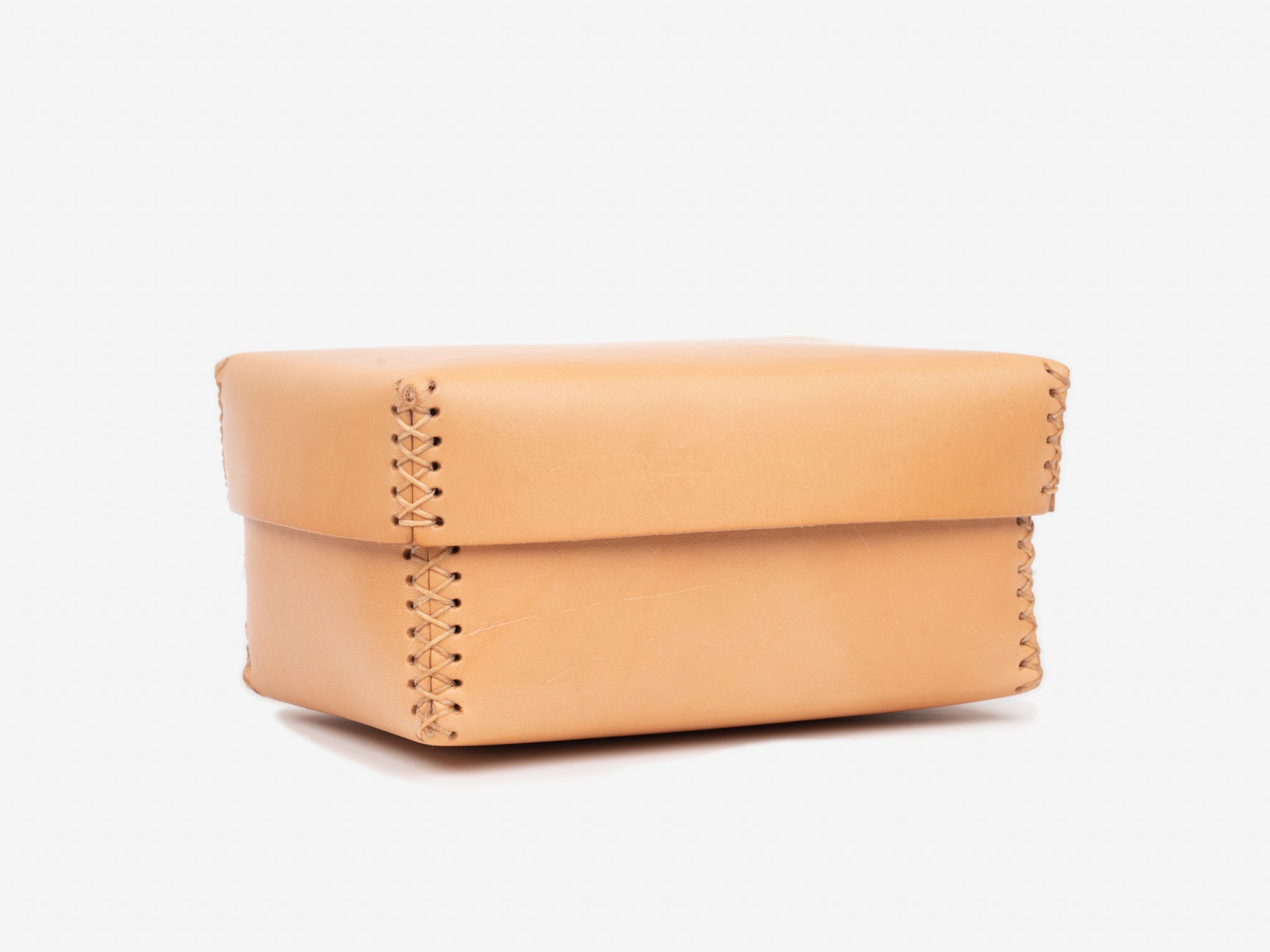 A small, natural leather box with a lid and visible hand-stitched seams along the edges, displayed on a plain white background.
