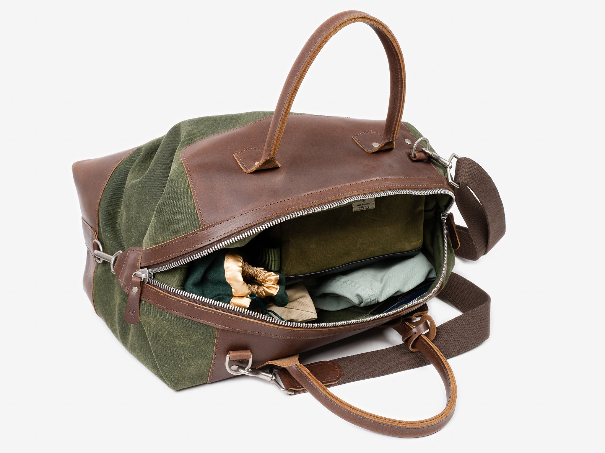 A top-down view of an open duffel bag with brown leather accents and green fabric sides, showing neatly packed clothing and a scrunchie inside.