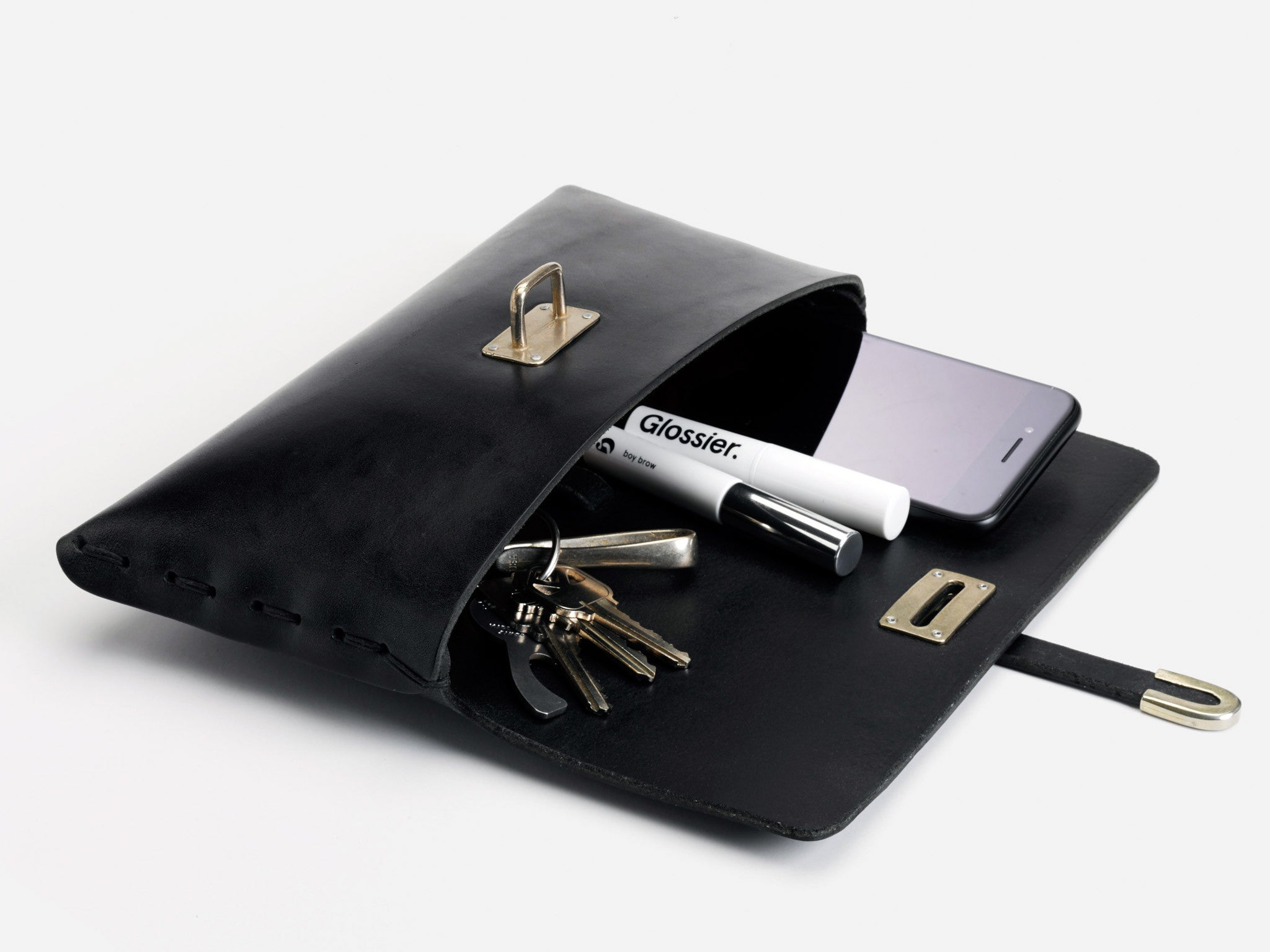 A black leather clutch lies open on a white surface, revealing a smartphone, several keys, and a Glossier-branded tube inside. The clasp and hardware are gold-colored.