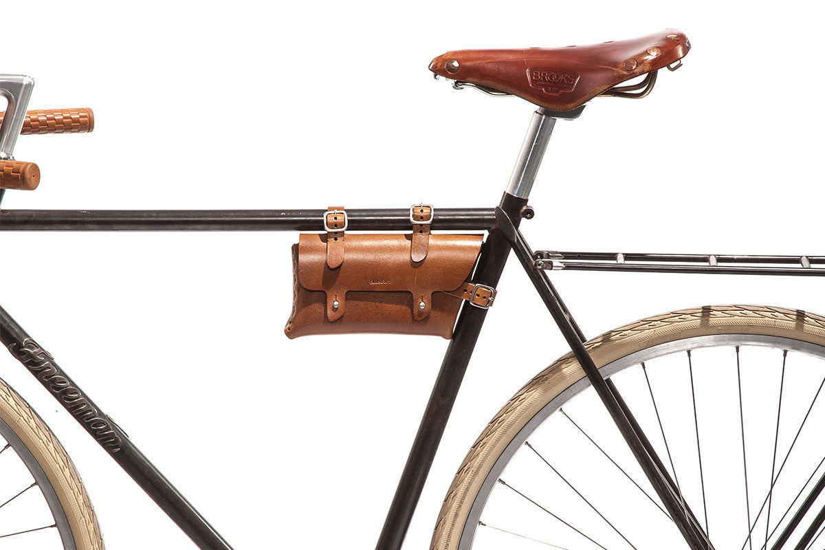 A close-up of a vintage-style bicycle with a brown leather saddle, matching handlebar grips, and a small brown leather tool bag attached to the frame above the rear wheel. The tire is cream-colored.