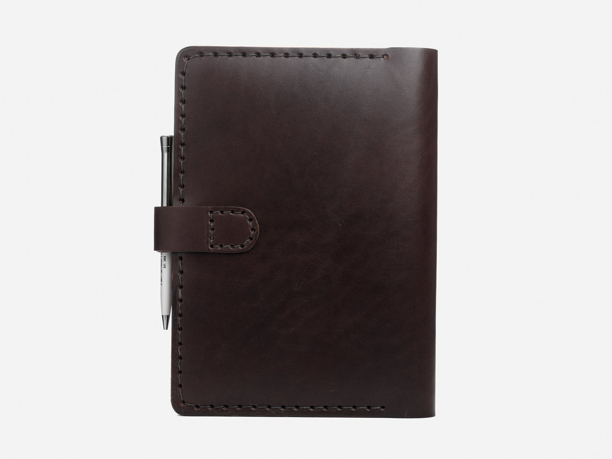A dark brown leather notebook with visible stitching, a flap closure, and a silver pen attached to the left side, shown against a plain white background.