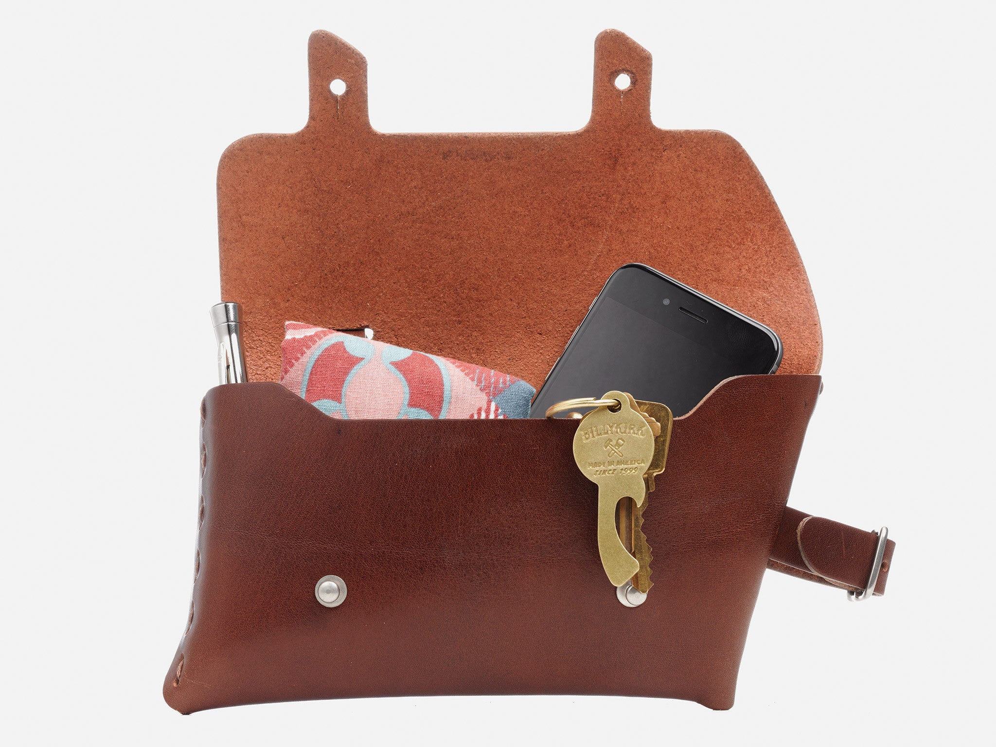 A brown leather pouch with its flap open, containing a smartphone, a pen, and a patterned cloth, with two brass keys hanging from a keyring on the front.